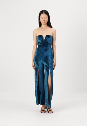 Combinaison en velours turquoise sans bretelles avec un décolleté en V plongeant, présentant des fentes latérales et une taille cintrée. Texture lisse et silhouette allongée.
