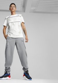 Hvid tekstureret t-shirt med grå sweatpants, der har sorte sideaccentuer og Puma-logo. Sorte, røde og blå atletiske sko fuldender outfittet.