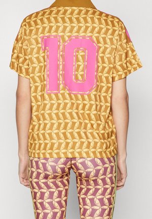 Personne portant un t-shirt jaune moutarde avec un motif de plumes et un grand numéro 10 rose dans le dos, associé à des leggings violets avec un motif similaire.