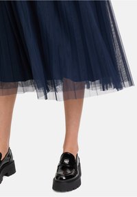 Gonna midi in tulle navy con tessuto trasparente a strati e orlo leggermente arricciato; abbinata a mocassini neri in vernice con fibbia decorativa.
