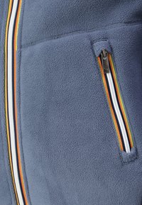 Fleece jacket in blue with a textured finish. Features a zippered pocket with multi-colored stripes: orange, yellow, white, and navy.