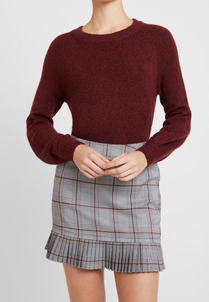 Vrouw draagt een bordeauxrode trui met lange mouwen en een grijze geruite minirok met geplooide zoom, staand tegen een effen achtergrond.