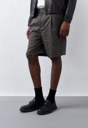 Man draagt donkergekleurde, patroonrijke kniebroeken, zwarte sokken, zwarte loafers, wit overhemd en zwart jasje tegen een effen achtergrond.