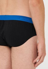 Zwarte katoenen onderbroeken met een blauwe elastische tailleband waarop het "Hugo"-logo staat, weergegeven vanuit de zijkant, waardoor de pasvorm en structuur zichtbaar zijn.