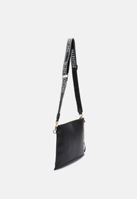 Černá kožená crossbody taška se zapínáním na zip, texturovaným povrchem, zlatými doplňky a nastavitelného popruhu s názvem značky v bílé barvě.