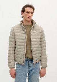 Light beige puffer jacket with horizontal quilted pattern, standing collar, front zipper, and a patch logo. Worn over a ribbed knit sweater.