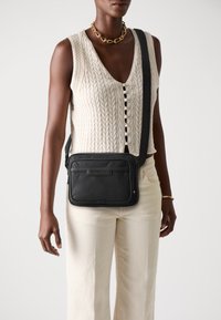 Schwarze Leder-Crossbody-Tasche mit rechteckiger Form, sichtbaren N�ähten und einem dezenten Logo-Detail. Ein einzelner verstellbarer Tragegurt für Komfort.