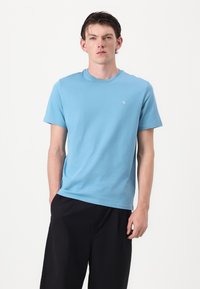 Ανοιχτό μπλε βαμβακερό μπλουζάκι με στρογγυλή λαιμόκοψη και κοντά μανίκια, διακοσμημένο με ένα μικρό λευκό λογότυπο στη αριστερή πλευρά του στήθους, συνδυασμένο με μαύρα σορτς.