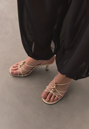 Piedi con sandali beige con tacco e cinturini, decorati con perline dorate, abbinati a pantaloni neri trasparenti a gamba larga su un pavimento grigio liscio.