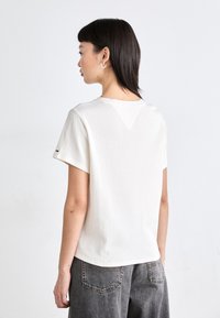 Camiseta de algodón blanca con ribetes, de mangas cortas y detalle en forma de V en la parte posterior, combinada con pantalones de mezclilla gris de cintura alta.
