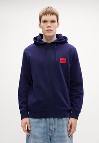 Marineblå zip-up hettegenser laget av myk stoff, med en rød rektangulær logo-patch og snorhette. Vanlig passform med ribbestrikkede mansjetter.
