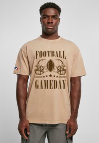 Beige bomulds t-shirt med "FOOTBALL GAMEDAY" grafik, en fodbold og to hjelme i mørkebrun. Løs pasform med korte ærmer.