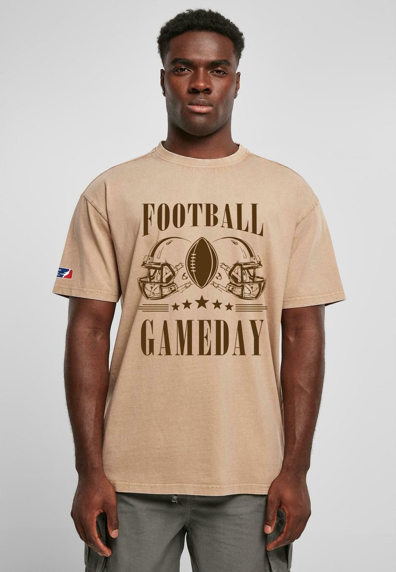 Beige bomulds t-shirt med "FOOTBALL GAMEDAY" grafik, en fodbold og to hjelme i mørkebrun. Løs pasform med korte ærmer.