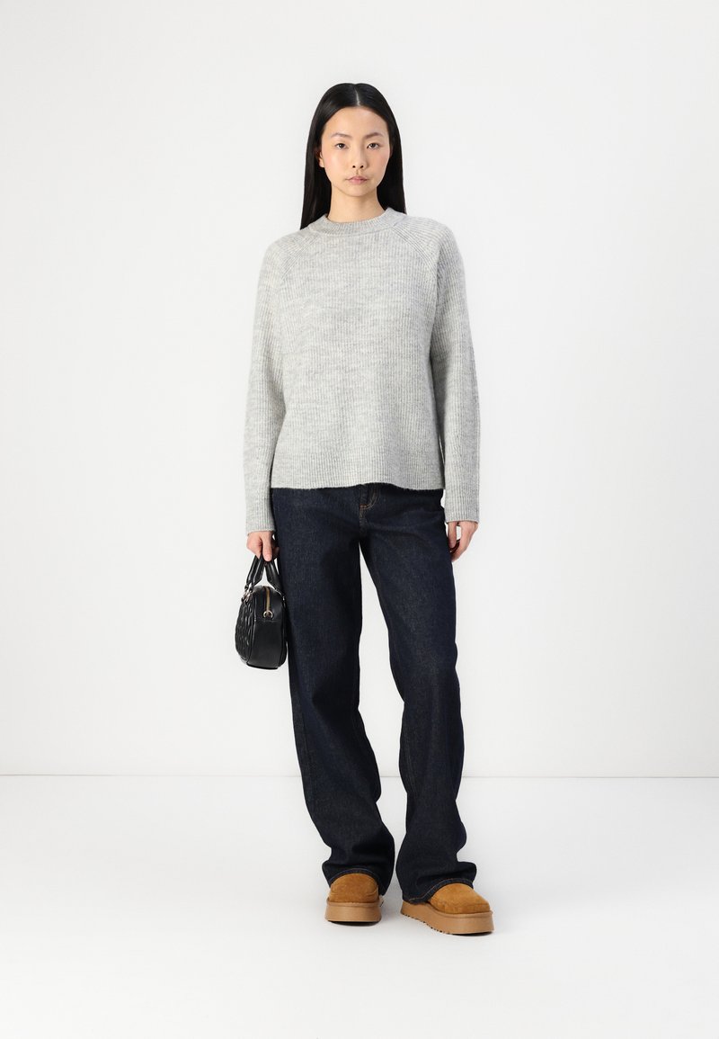Hellgrauer Strickpullover mit lockerer Passform, dunkelblaue Jeans und beigefarbene Plateauschuhe. Schwarze Handtasche mit strukturiertem Oberflächenmaterial.