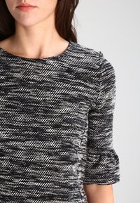 Haut texturé noir et blanc avec un col rond, des manches trois-quarts et un tissu tissé présentant un motif moucheté.