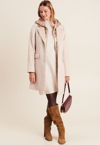 Manteau en laine beige avec capuche, superposé à une robe pull crème. Des bottes en daim marron jusqu'aux genoux et un sac à bandoulière bordeaux complètent le look.