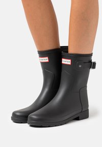 Botas de lluvia de goma negras con punta redondeada, de altura hasta la mitad de la pantorrilla, que cuentan con un cinturón lateral, suela texturizada y etiqueta de marca roja y blanca.