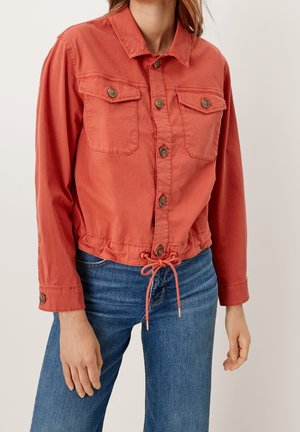 Femme portant une veste couleur rouille avec fermeture boutonnée, taille avec cordon de serrage et deux poches poitrine, associée à un jean bleu.