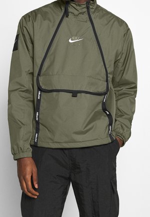 Mężczyzna noszący oliwkowo-zieloną kurtkę przeciwwiatrową Nike Air z przednią kieszenią z klapką na zamek błyskawiczny i czarnym napisem Nike Air na ramiączkach, w połączeniu z czarnymi spodniami.