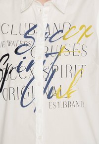 Camicia bianca con bottoni caratterizzata da testo e design grafici audaci e multicolori in blu, giallo e nero. Tessuto in cotone morbido con una texture liscia.