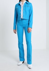 Veste en denim bleu clair courte avec des boutons argentés et deux poches à rabat, associée à un pantalon ajusté assorti et des bottes blanches à bout pointu.