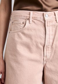 Pantalones de mezclilla rosa claro con cintura alta, diseño de cinco bolsillos, botones metálicos y detalles cosidos. Textura suave con un ligero brillo.