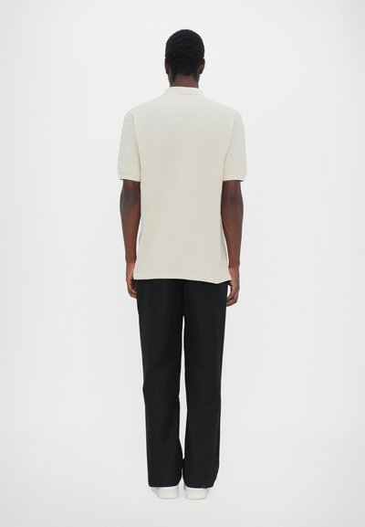Polo beige avec une texture douce, à manches courtes et col, associé à un pantalon noir ample et des chaussures blanches.