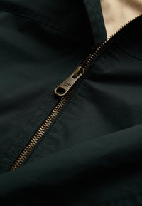 Chaqueta de tela verde oscuro con una cremallera de color dorado. La textura parece lisa, con costuras visibles a lo largo de los bordes.