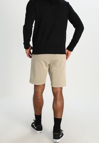 Beige šortky s mírně texturovanou tkaninou, v kombinaci s černou mikinou. Model má na sobě černé tenisky a ponožky, zobrazeno ze zadního pohledu.