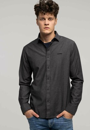 Młody mężczyzna z kręconymi włosami w ciemnoszarej koszuli zapinanej na guziki i niebieskich dżinsach, stojący z rękami w kieszeniach na jednolitym tle.