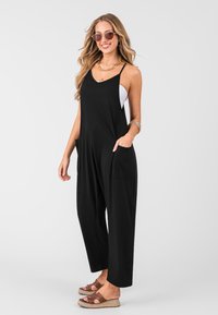 Jumpsuit nero senza maniche realizzato in morbido tessuto, caratterizzato da una vestibilità ampia, tasche laterali e fondo a risvolto. Indossato con sandali espadrilles marroni.