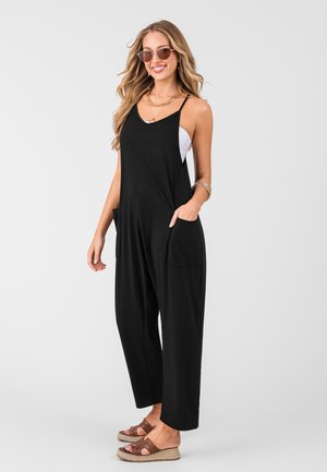Combinaison noire sans manches en tissu doux, avec une coupe décontractée, des poches latérales et un bas à ourlet. Portée avec des sandales en espadrilles marron.