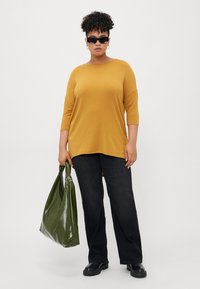 Person wearing black sunglasses, mustard yellow long-sleeve top, black pants, black boots, holding large olive green handbag.
