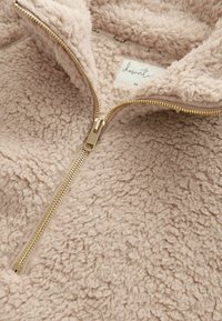 Fleece-pullover i blød beige med en tekstureret, blød overflade. Har en gulddetaljeret lynlås og en label inde i kraven.