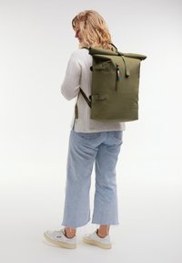 Zaino verde con top arrotolabile, spallacci regolabili, tasca frontale con zip e accenti di colore a contrasto. Materiale liscio e resistente con design minimalista.