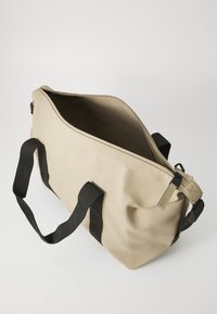 Μπεζ αδιάβροχη τσάντα με μαύρα λουριά, ευρύχωρο κύριο διαμέρισμα και κλείσιμο με φερμουάρ στην κορυφή. Ομαλή υφή και μινιμαλιστικός σχεδιασμός.