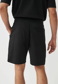 Bermudas negras fabricadas en una mezcla de algodón, con una cinturilla ajustable con cordón, bolsillos laterales y un bolsillo trasero único con detalle de solapa.