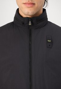 Black jacket with high collar, full zipper, and branded patch. Features a lightweight, textured material with a clean, minimalist design.