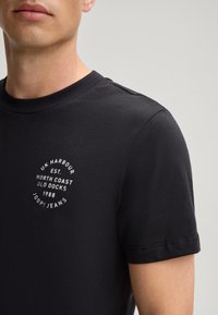 T-shirt noir à manches courtes en coton doux, avec un graphique circulaire blanc sur la poitrine comportant des détails textuels et un col rond.