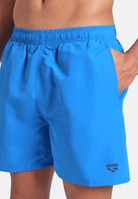 Uomo che indossa pantaloni da bagno blu brillante con cintura elastica, mano nella tasca, e logo "arena" vicino all'orlo della gamba destra.