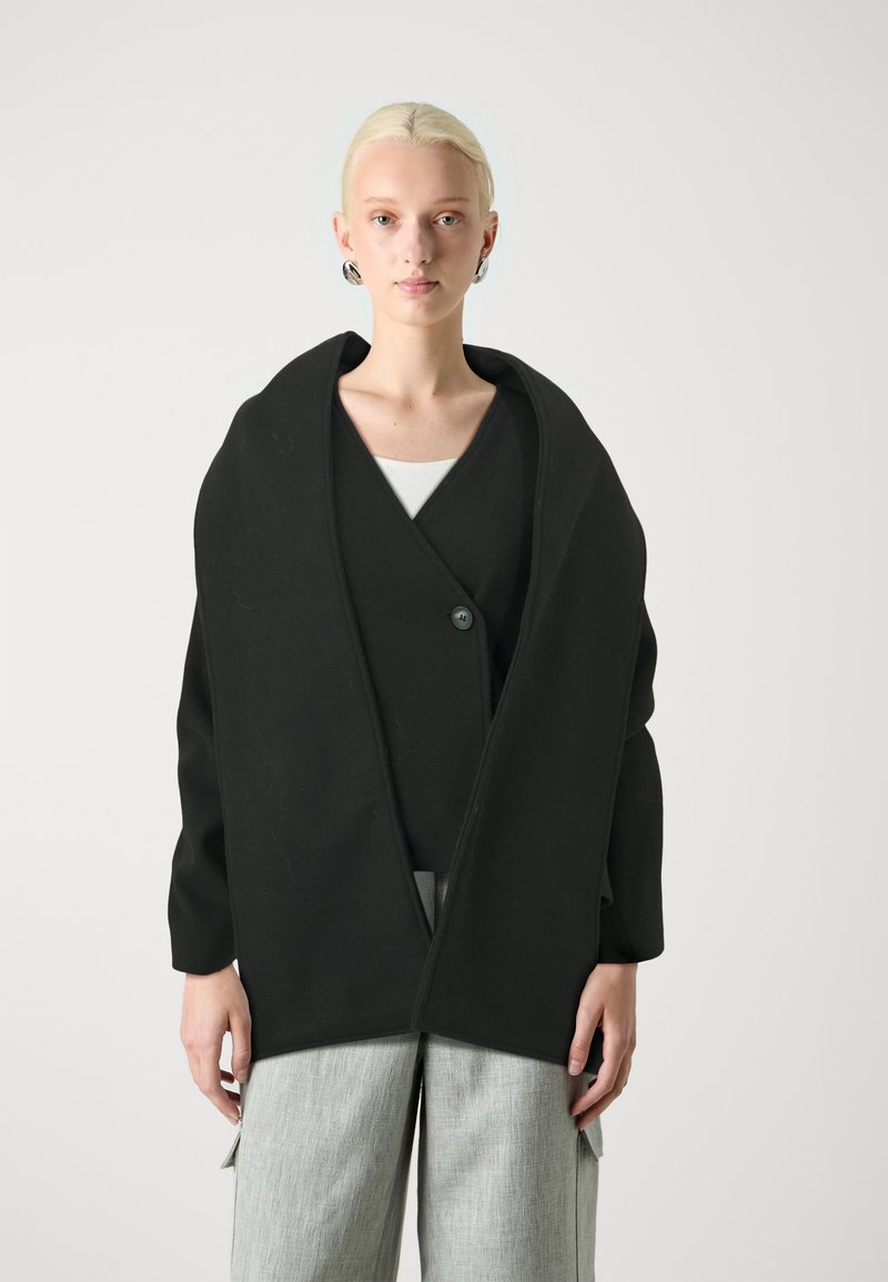 Schwarze Wickel-Cardigan mit einem einzigen Knopfverschluss, aus weichem Stoff gefertigt, verfügt über einen breiten Kragen und eine schlichte Textur, getragen über einem weißen Oberteil.