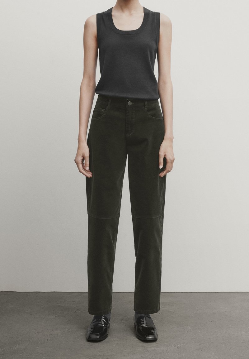 Personne portant un haut noir sans manches, un pantalon sombre taille haute avec un détail de couture, et des chaussures noires vernies, debout contre un mur clair uni.