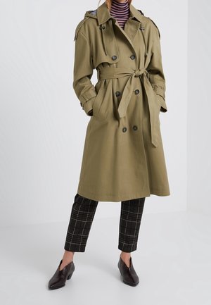 Femme portant un trench-coat croisé vert olive avec ceinture, un col roulé rayé, un pantalon court à carreaux et des chaussures sombres à bout pointu, debout.