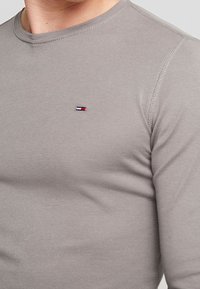 Graues Langarm-T-Shirt aus weichem Stoff, mit rundem Ausschnitt und einem kleinen Logotag in Rot, Weiß und Blau auf der Brust.