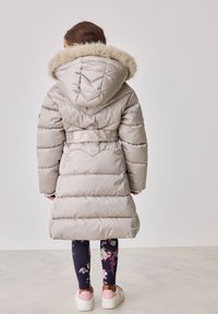 Beige színű pufikabát, egy szőrmebéléssel ellátott kapucnival, gumírozott derékrésszel és gradiens varrással a háton. Virágmintás leggings-szel és tornacipővel viselve.