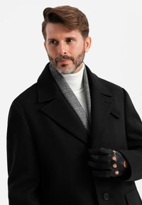 Abrigo de lana negro con solapas, superpuesto a un suéter de cuello alto blanco. Una bufanda a cuadros está drapeada, y se llevan guantes de cuero negro con cortes.