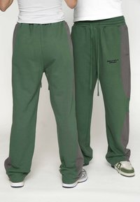 Pantaloni de trening verzi cu panouri laterale gri, talie cu șnur și un logo pe șoldul drept. Materialul pare moale și confortabil.