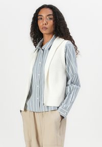Gilet tricoté texturé, blanc cassé, par-dessus une chemise à rayures bleu clair à boutons. Comprend un col et une fermeture éclair ; associé à un pantalon beige.