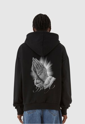 Zwarte hoodie met een grafisch ontwerp van samengevouwen handen en een rozenkrans op de achterkant, met een halo-effect en het woord "Blessed" in sierlijke letters.