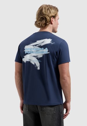 Man met blond haar die een marineblauw T-shirt draagt met witte verfstrepen en lichtblauwe tekst "RECORDS & PUBLISHING" op de achterkant.